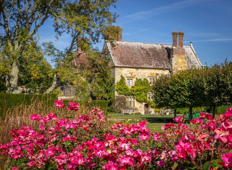 Historic stone house framed by bright pink flowers and mature trees, set within a well-kept formal garden.